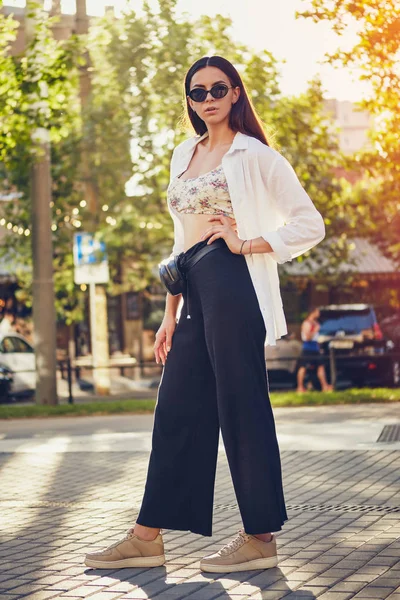 Retrato de uma menina de óculos escuros posando na cidade. Vestido em cima com estampa floral, camisa branca, calças pretas, bolsa de cintura e tênis . — Fotografia de Stock