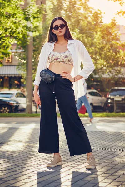 Retrato de uma menina de óculos escuros posando na cidade. Vestido em cima com estampa floral, camisa branca, calças pretas, bolsa de cintura e tênis . — Fotografia de Stock