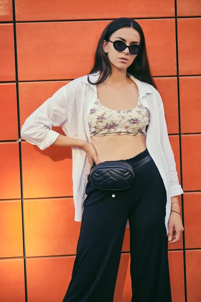 Portrait of girl in dark sunglasses posing in city against orange building. Dressed in top with floral print, white shirt, black trousers, waist bag. – stockfoto