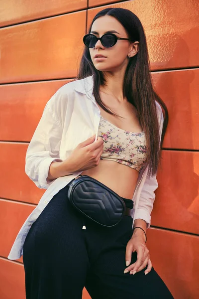Portrait of girl in dark sunglasses posing in city against orange building. Dressed in top with floral print, white shirt, black trousers, waist bag. – stockfoto