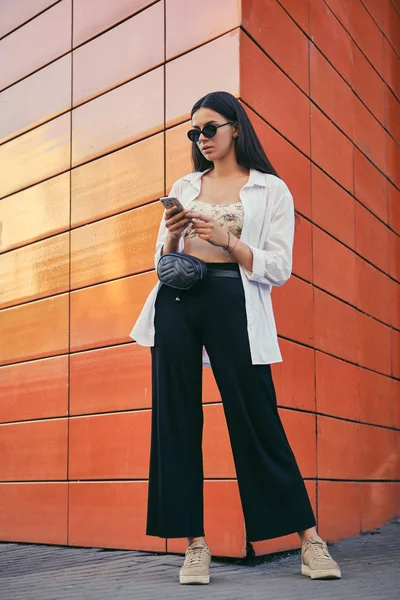Girl in sunglasses posing in city against an orange building. Dressed in top with floral print, white shirt, black trousers, waist bag and sneakers. – stockfoto