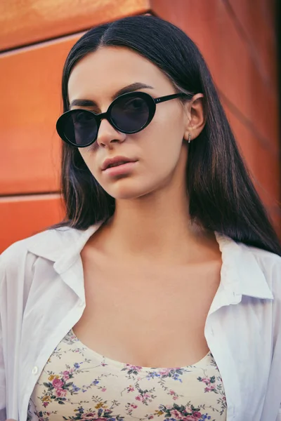 Egy lány portréja sötét napszemüvegben, aki a városban pózol a narancssárga épület ellen. Fent virágmintás ruhában, fehér ingben.. — Stock Fotó