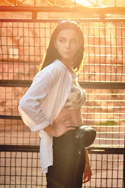 Retrato de una chica con gafas de sol oscuras posando en la ciudad contra una valla entrelazada. Vestido en la parte superior con estampado floral, camisa blanca, pantalones negros, bolso de cintura . —  Fotos de Stock