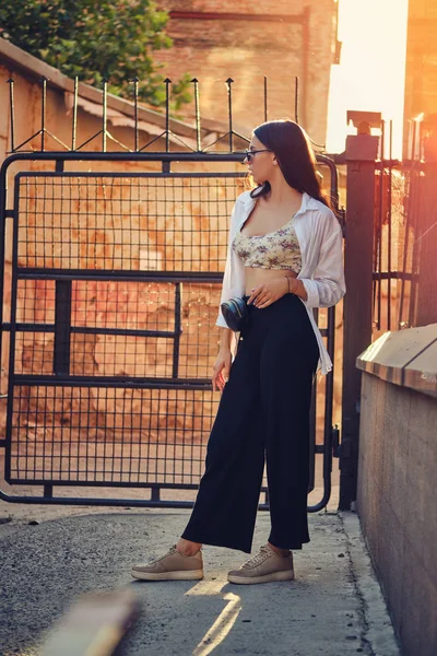 Porträt eines Mädchens mit dunkler Sonnenbrille, das in der Stadt posiert. Bekleidet mit Oberteil mit Blumenmuster, weißem Hemd, schwarzer Hose, Taille und Turnschuhen. — Stockfoto