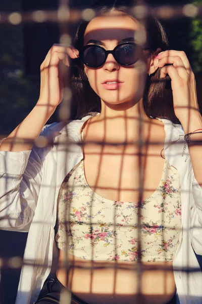 Retrato de niña en gafas de sol posando en la ciudad detrás de una valla trellised. Vestido en la parte superior con estampado floral, camisa blanca, pantalones negros, bolso de cintura . —  Fotos de Stock