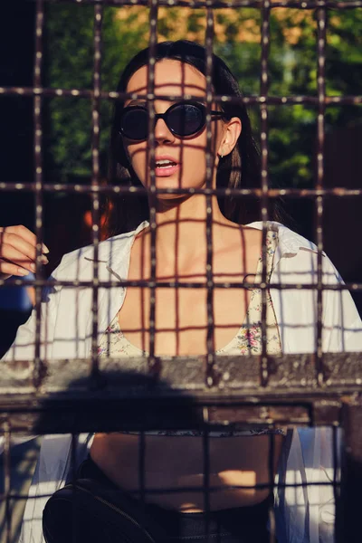 Portret van een meisje met een zonnebril in de stad achter een tralies. Bovenkant gekleed met bloemenprint, wit shirt, zwarte broek, taille tas. — Stockfoto