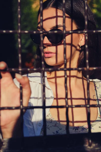 Portret van een meisje met een zonnebril in de stad achter een tralies. Bovenkant gekleed met bloemenprint, wit shirt, zwarte broek, taille tas. — Stockfoto