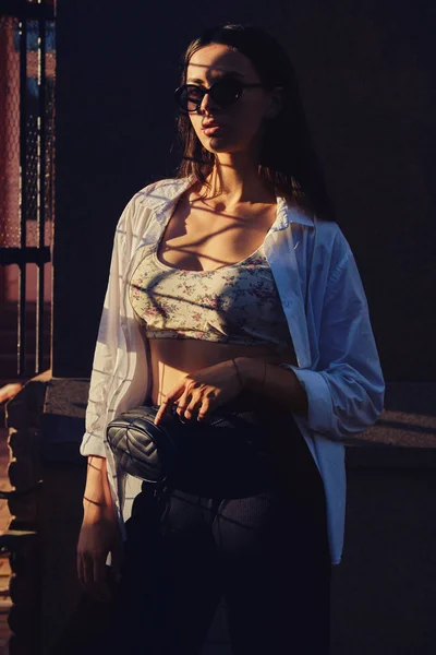 Portret van een meisje in een donkere zonnebril die poseert in de stad vlakbij een tralies hek. Bovenkant gekleed met bloemenprint, wit shirt, zwarte broek, taille tas. — Stockfoto