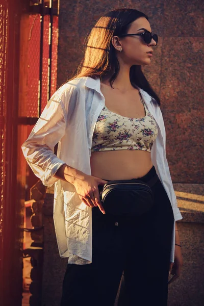 Retrato de uma menina em óculos escuros posando na cidade perto de cerca trellised. Vestido em cima com estampa floral, camisa branca, calças pretas, bolsa de cintura. — Fotografia de Stock