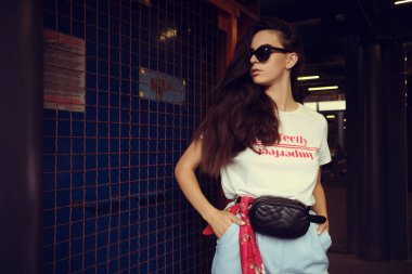 Koyu güneş gözlüklü bir kızın portresi alışveriş merkezinin yanındaki mavi kapıda poz veriyor. Beyaz tişört, mavi pantolon, siyah bel çantası, kırmızı mendil..