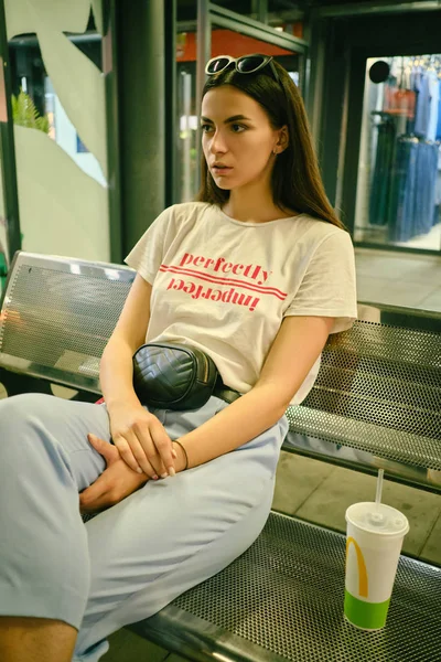 Retrato de menina com óculos escuros na cabeça posando sentado no banco no shopping center. Vestido em t-shirt branca, calças azuis, bolsa de cintura preta. — Fotografia de Stock