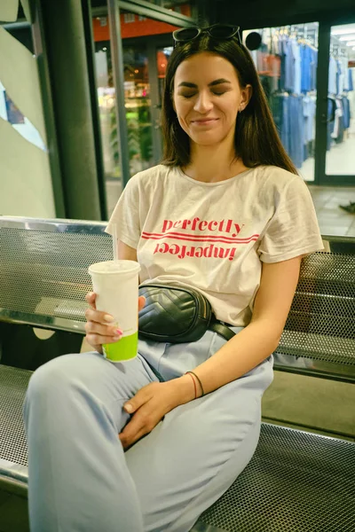 Retrato de menina com óculos escuros na cabeça posando sentado no banco no shopping center. Vestido em t-shirt branca, calças azuis, bolsa de cintura preta. — Fotografia de Stock