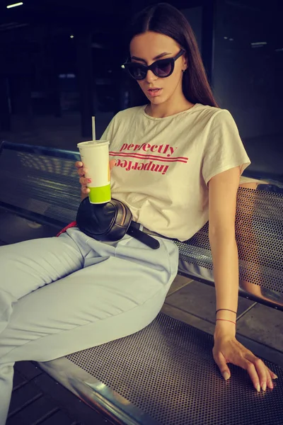 Retrato de menina em óculos escuros posando sentado no banco no shopping center. Vestido em t-shirt branca, calças azuis, bolsa de cintura preta. — Fotografia de Stock