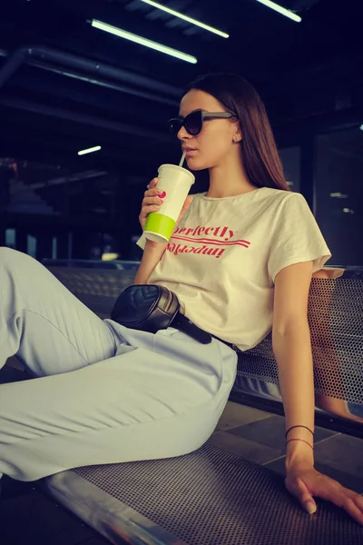 Ritratto di ragazza in occhiali da sole scuri in posa seduto sulla panchina nel centro commerciale. Vestito con t-shirt bianca, pantaloni blu, borsa in vita nera. — Foto Stock