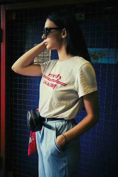 Retrato de uma menina em óculos escuros posando em um portão azul perto do shopping. Vestido em t-shirt branca, calças azuis, saco da cintura preta, lenço vermelho. — Fotografia de Stock