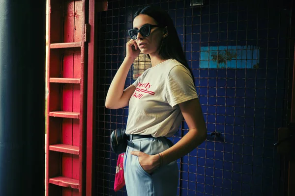 Porträt eines Mädchens mit dunkler Sonnenbrille, das vor einem blauen Tor in der Nähe des Einkaufszentrums posiert. Bekleidet mit weißem T-Shirt, blauer Hose, schwarzer Taille, rotem Halstuch. — Stockfoto