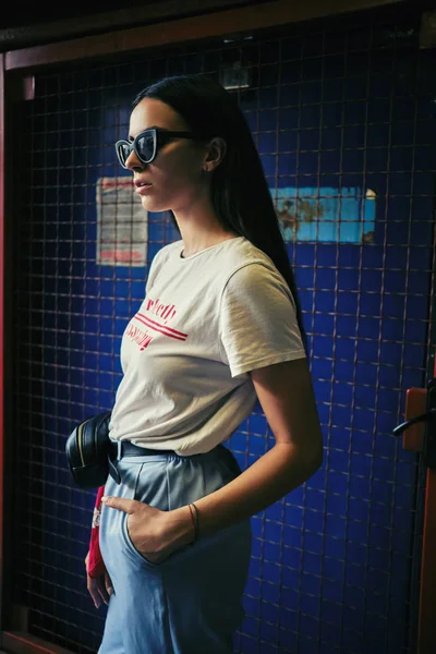 Retrato de uma menina em óculos escuros posando em um portão azul perto do shopping. Vestido em t-shirt branca, calças azuis, saco da cintura preta, lenço vermelho. — Fotografia de Stock