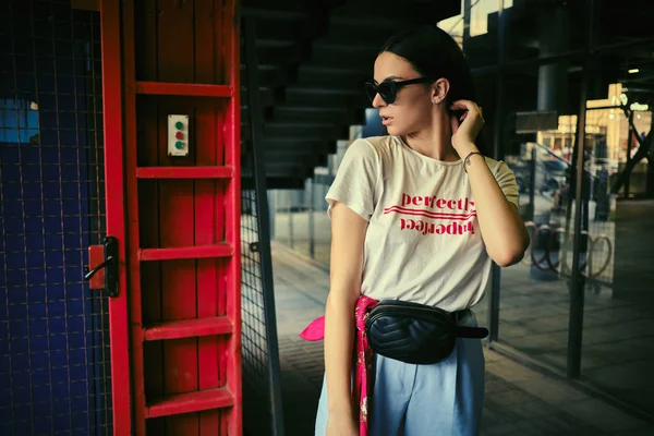 Porträt eines Mädchens mit dunkler Sonnenbrille, das vor einem blauen Tor in der Nähe des Einkaufszentrums posiert. Bekleidet mit weißem T-Shirt, blauer Hose, schwarzer Taille, rotem Halstuch. — Stockfoto