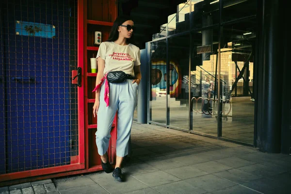 Porträt eines Mädchens mit dunkler Sonnenbrille, das vor einem blauen Tor in der Nähe des Einkaufszentrums posiert. Bekleidet mit weißem T-Shirt, blauer Hose, schwarzer Taille, Turnschuhen. — Stockfoto