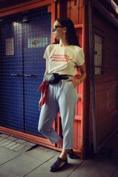 Retrato de uma menina em óculos escuros posando em um portão azul perto do shopping. Vestido em t-shirt branca, calças azuis, bolsa de cintura preta, tênis. — Fotografia de Stock