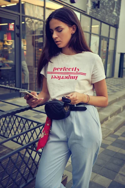 Retrato de uma menina posando perto de um shopping da cidade, usando smartphone. Vestido em t-shirt branca, calças azuis, saco da cintura preta, lenço vermelho. — Fotografia de Stock