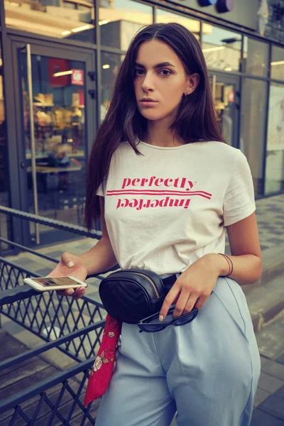 Retrato de uma menina posando perto de um shopping da cidade, usando smartphone. Vestido em t-shirt branca, calças azuis, saco da cintura preta, lenço vermelho. — Fotografia de Stock