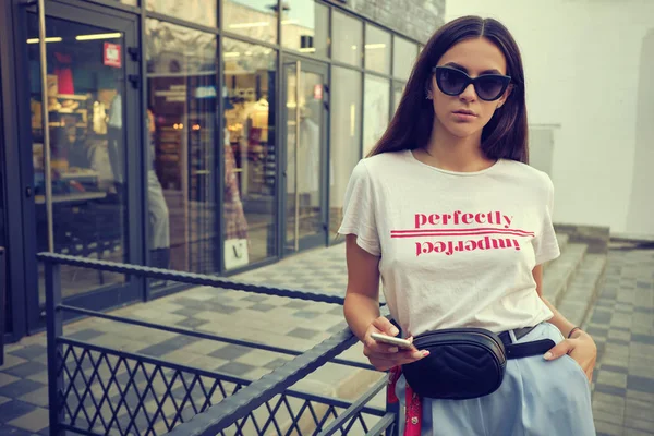 Портрет девушки в солнцезащитных очках, позирующей у городской черты с помощью смартфона. Был одет в белую футболку, синие брюки, черную сумку до пояса, красный платок. — стоковое фото