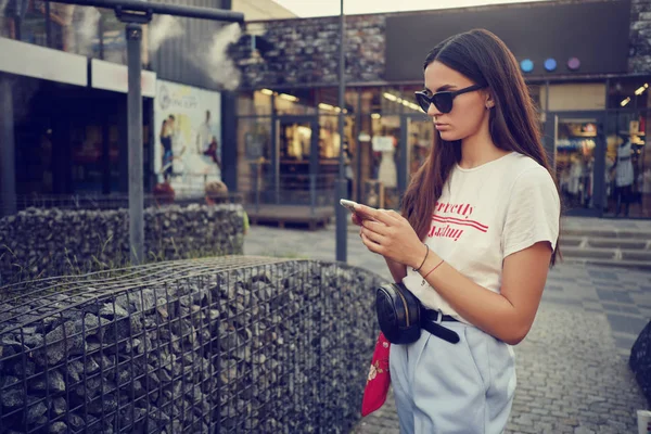 Porträt eines Mädchens mit Sonnenbrille, das in der Nähe eines Einkaufszentrums mit einem Smartphone posiert. Bekleidet mit weißem T-Shirt, blauer Hose, schwarzer Taille, rotem Halstuch. — Stockfoto