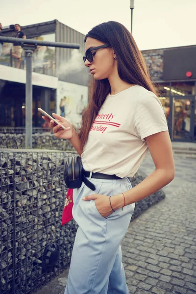 Porträt eines Mädchens mit Sonnenbrille, das in der Nähe eines Einkaufszentrums mit einem Smartphone posiert. Bekleidet mit weißem T-Shirt, blauer Hose, schwarzer Taille, rotem Halstuch. — Stockfoto