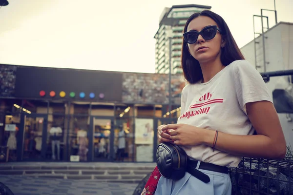 Porträt eines Mädchens mit dunkler Sonnenbrille, das in der Nähe eines Einkaufszentrums posiert. Bekleidet mit weißem T-Shirt, blauer Hose, schwarzer Taille, rotem Halstuch. — Stockfoto