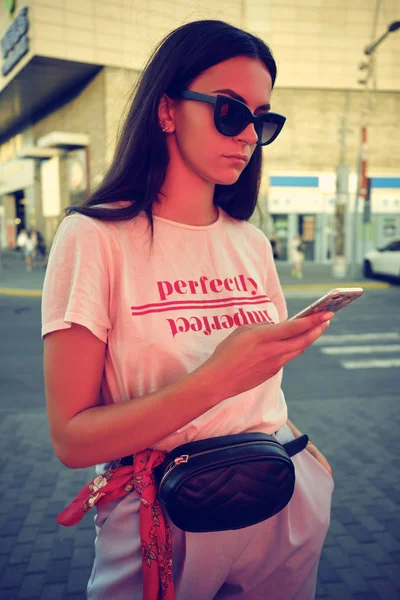 Retrato de uma menina em óculos de sol posando perto de um shopping da cidade, usando smartphone. Vestido em t-shirt branca, calças azuis, saco da cintura preta, lenço vermelho. — Fotografia de Stock