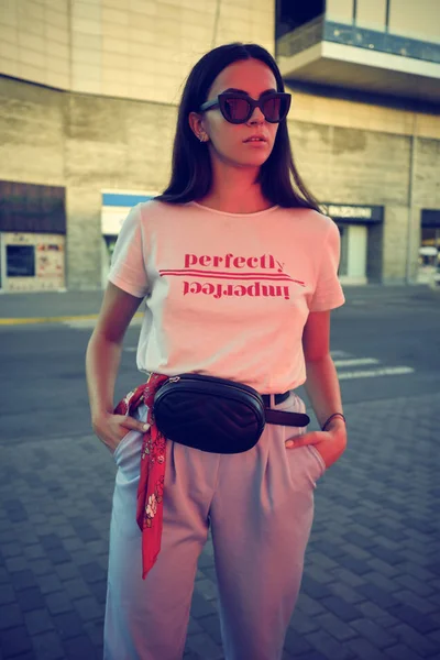 Retrato de uma menina em óculos escuros posando perto de um shopping da cidade. Vestido em t-shirt branca, calças azuis, saco da cintura preta, lenço vermelho. — Fotografia de Stock