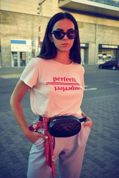 Retrato de uma menina em óculos escuros posando perto de um shopping da cidade. Vestido em t-shirt branca, calças azuis, saco da cintura preta, lenço vermelho. — Fotografia de Stock