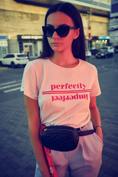 Retrato de una niña en gafas de sol oscuras que representa cerca de un centro comercial de la ciudad. Vestido con camiseta blanca, pantalones azules, bolsa de cintura negra, pañuelo rojo.. —  Fotos de Stock