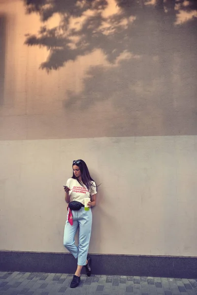 Retrato de uma menina em óculos escuros posando na cidade, contra uma parede. Vestido em t-shirt branca, calças azuis, bolsa de cintura preta e tênis. — Fotografia de Stock