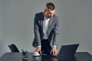 Klasik takım elbiseli, şık bıyıklı sakallı iş adamı, masada gri arka planda otururken dizüstü bilgisayarda çalışıyor..