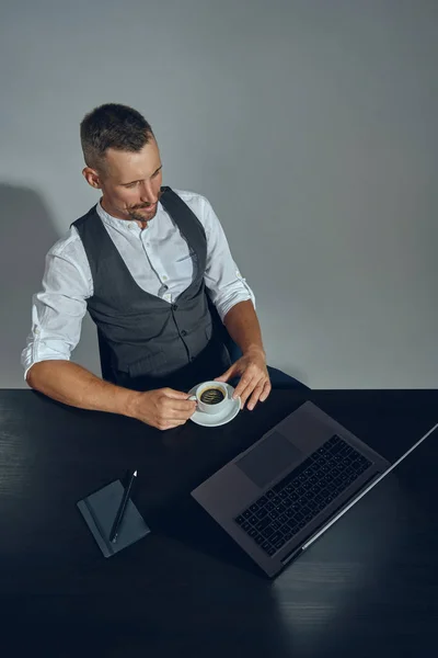 Szakállas üzletember elegáns bajusszal, klasszikus öltönyben nézi a laptopot, miközben az asztalnál ül az irodában, szürke háttérrel. Felülnézet. — Stock Fotó