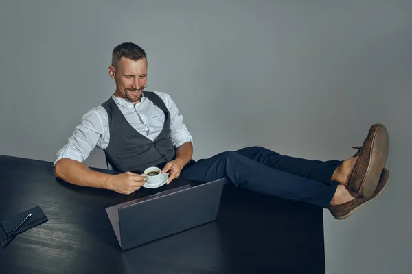 Der bärtige Geschäftsmann mit dem stylischen Schnurrbart, im klassischen Anzug, hat seine Beine auf den Tisch gestellt, eine Tasse Kaffee in der Hand, schaut auf den Laptop. Ansicht von oben. — Stockfoto
