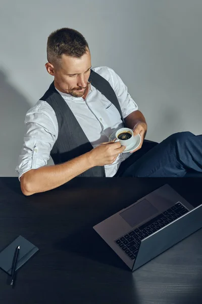 Der bärtige Geschäftsmann mit dem stylischen Schnurrbart und dem klassischen Anzug blickt auf den Laptop, während er im Büro am Tisch sitzt und Kaffee trinkt. Ansicht von oben. — Stockfoto