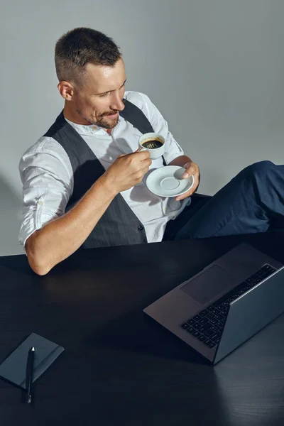 Der bärtige Geschäftsmann mit dem stylischen Schnurrbart und dem klassischen Anzug blickt auf den Laptop, während er im Büro am Tisch sitzt und Kaffee trinkt. Ansicht von oben. — Stockfoto