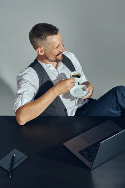 Бизнесмен со стильной гримасой, одетый в классический костюм, улыбается, отводя взгляд, сидя за столом в офисе и выпивая кофе. Вид сверху. — стоковое фото