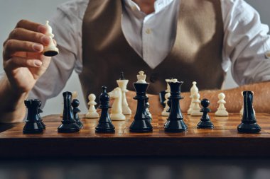 Klasik kahverengi yelek ve beyaz gömlek giymiş bir adamın elleri masada oturuyor ve tek başına satranç oynuyor. Gri arkaplan, yakın çekim.