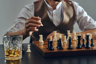 Kahverengi yelek giymiş, satranç oynayan beyaz gömlekli bir adamın elleri. Yakınlarda bir bardak viski var. Gri arkaplan, yakın plan.