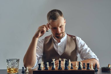 Kahverengi yelek giymiş şık bıyıklı bir adam, beyaz gömlek satranç oynuyor. Yakınlarda bir bardak viski, kül tablası, puro. Gri arkaplan, yakın plan.