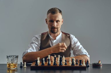 Kahverengi yelek giymiş şık bıyıklı bir adam, beyaz gömlek satranç oynuyor. Yakınlarda bir bardak viski, kül tablası, puro. Gri arkaplan, yakın plan.