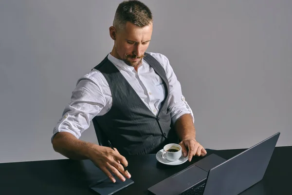 Szakállas üzletember elegáns bajusszal, klasszikus öltönyben dolgozik a laptopnál, miközben az asztalnál ül az irodában, szürke háttér. — Stock Fotó