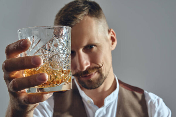 Man with stylish mustache, dressed in classic brown vest, white shirt is sitting at the table, enjoying whiskey. Grey background, close-up shot.