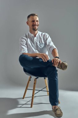 Klasik beyaz gömlekli ve mavi pantolonlu şık bıyıklı adam sandalyede oturuyor. Gri arkaplan, yakın çekim.