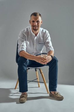 Klasik beyaz tişörtlü ve mavi pantolonlu şık bıyıklı bir adam sandalyeye oturmuş, elinde bir bardak viski tutuyor. Gri arkaplan, yakın çekim.