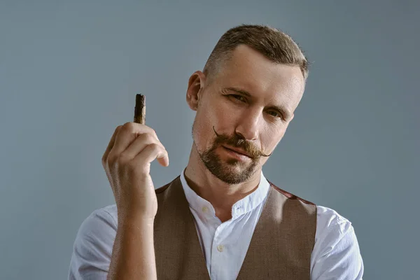 Un homme avec une moustache élégante, vêtu d'une veste brune et d'une chemise blanche pose assis sur une table, dégustant un cigare. Fond gris, gros plan. — Photo